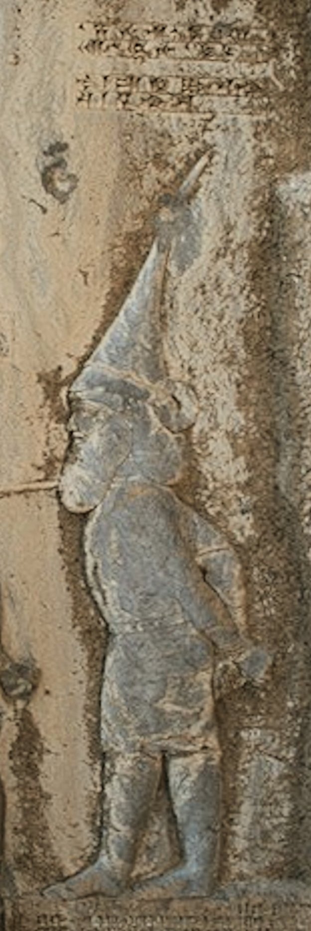 Carved figure of the rebel Skunkha wearing a tall pointed hat, roped at the neck, at the end of the line of captives on the Behistun relief