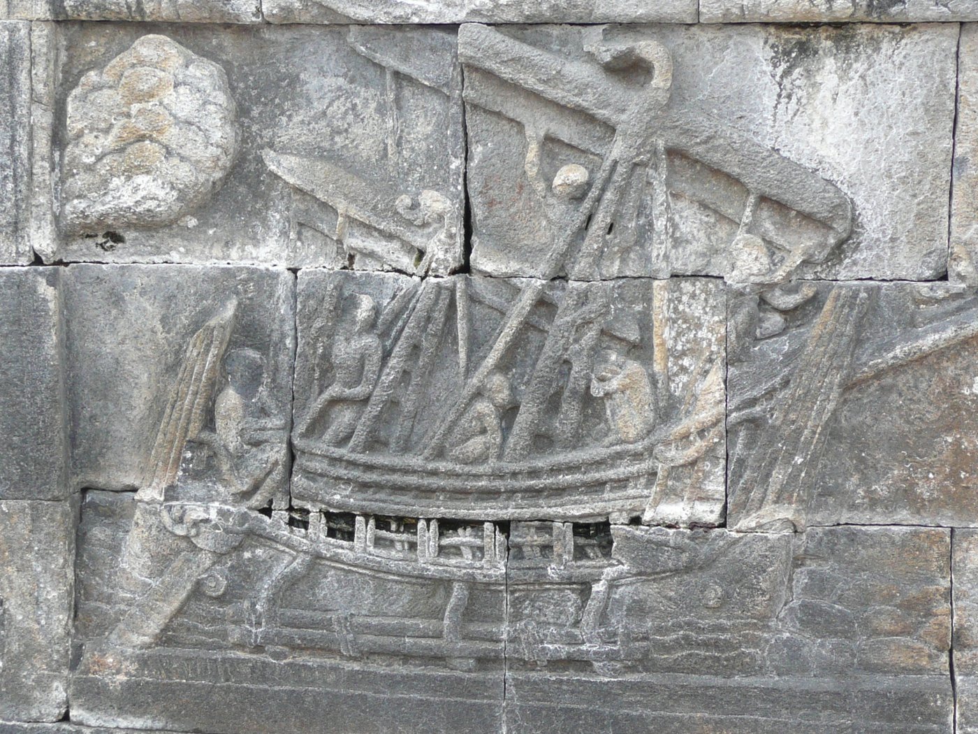 A stone relief at Borobudur depicting a large ancient sailing ship with outriggers and billowing sails crewed by several figures