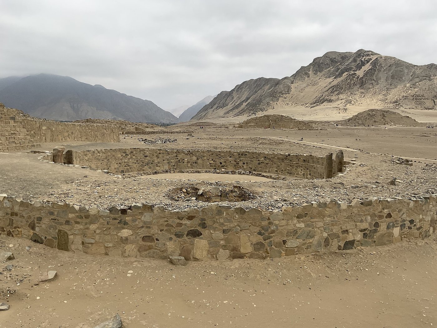 A sunken circular stone plaza at Caral, set into the ground in front of a large pyramid mound
