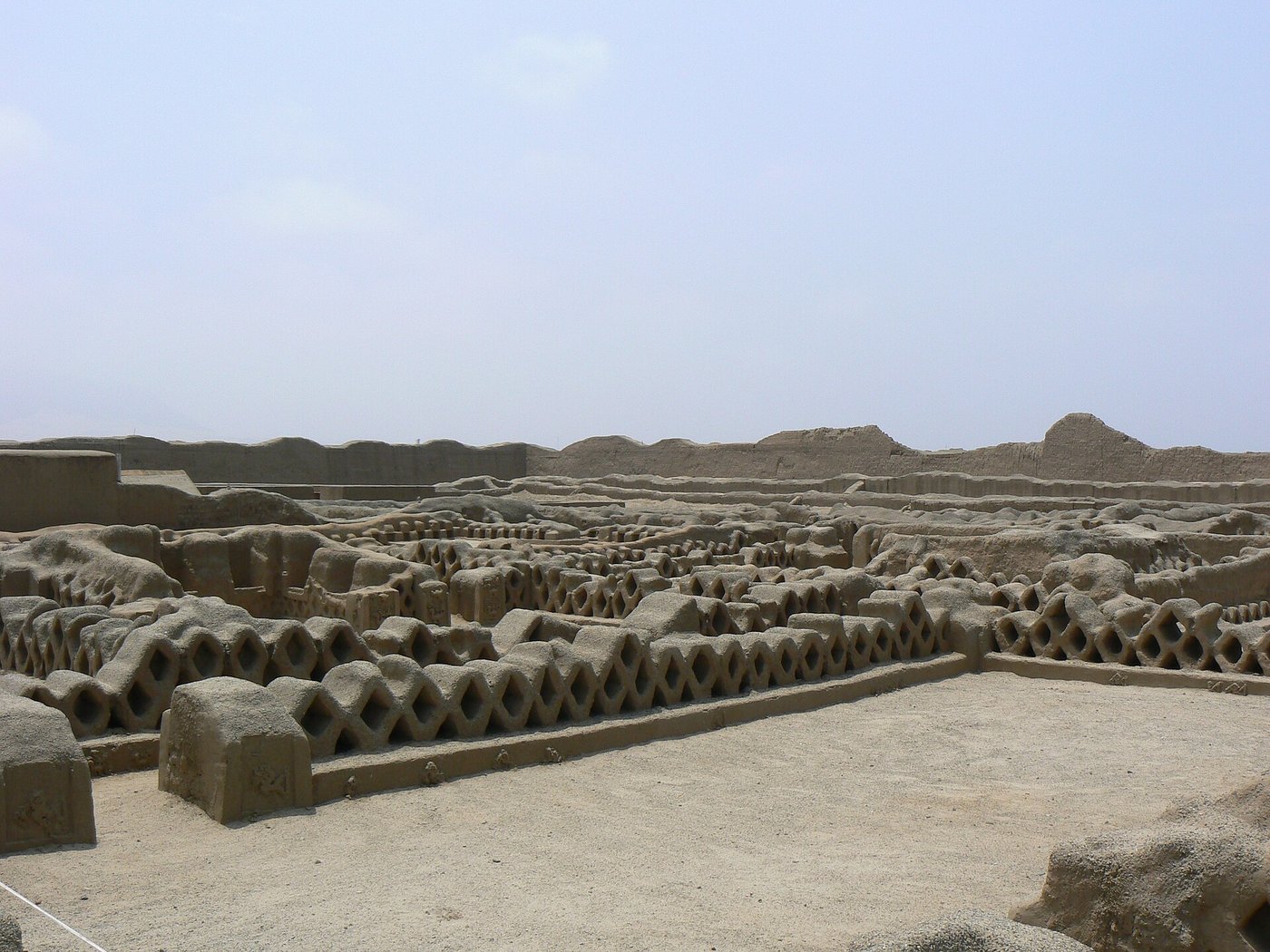 A corridor of adobe walls pierced with a diamond lattice resembling a fishing net, lining rows of small storerooms