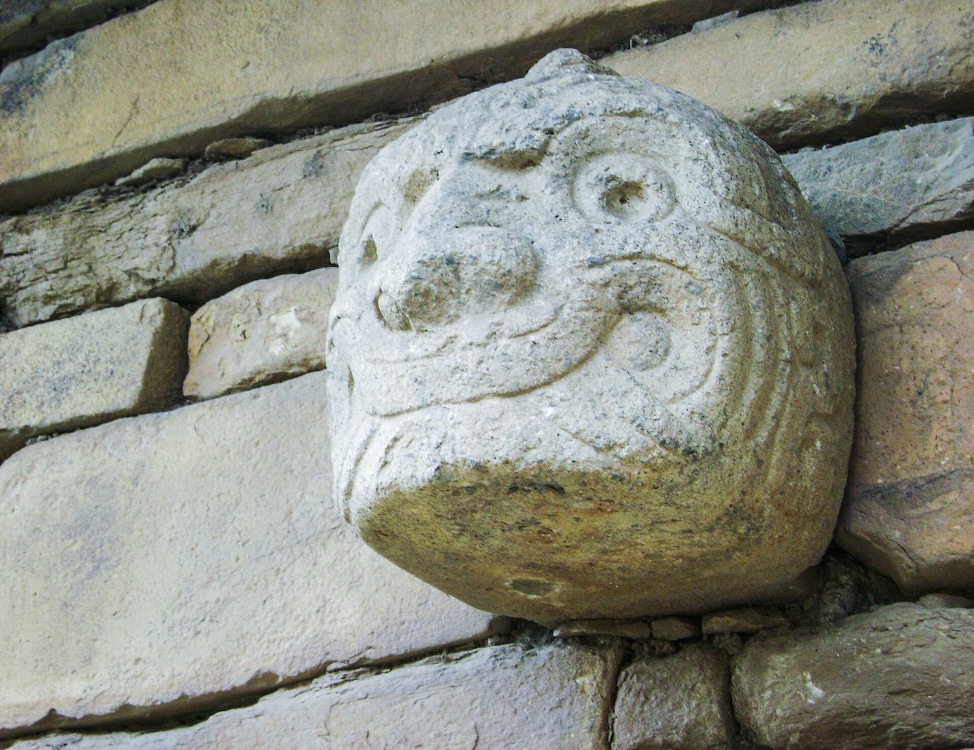A stone tenon head still set in the outer wall at Chavín de Huántar, a carved face with bulging features projecting from the masonry