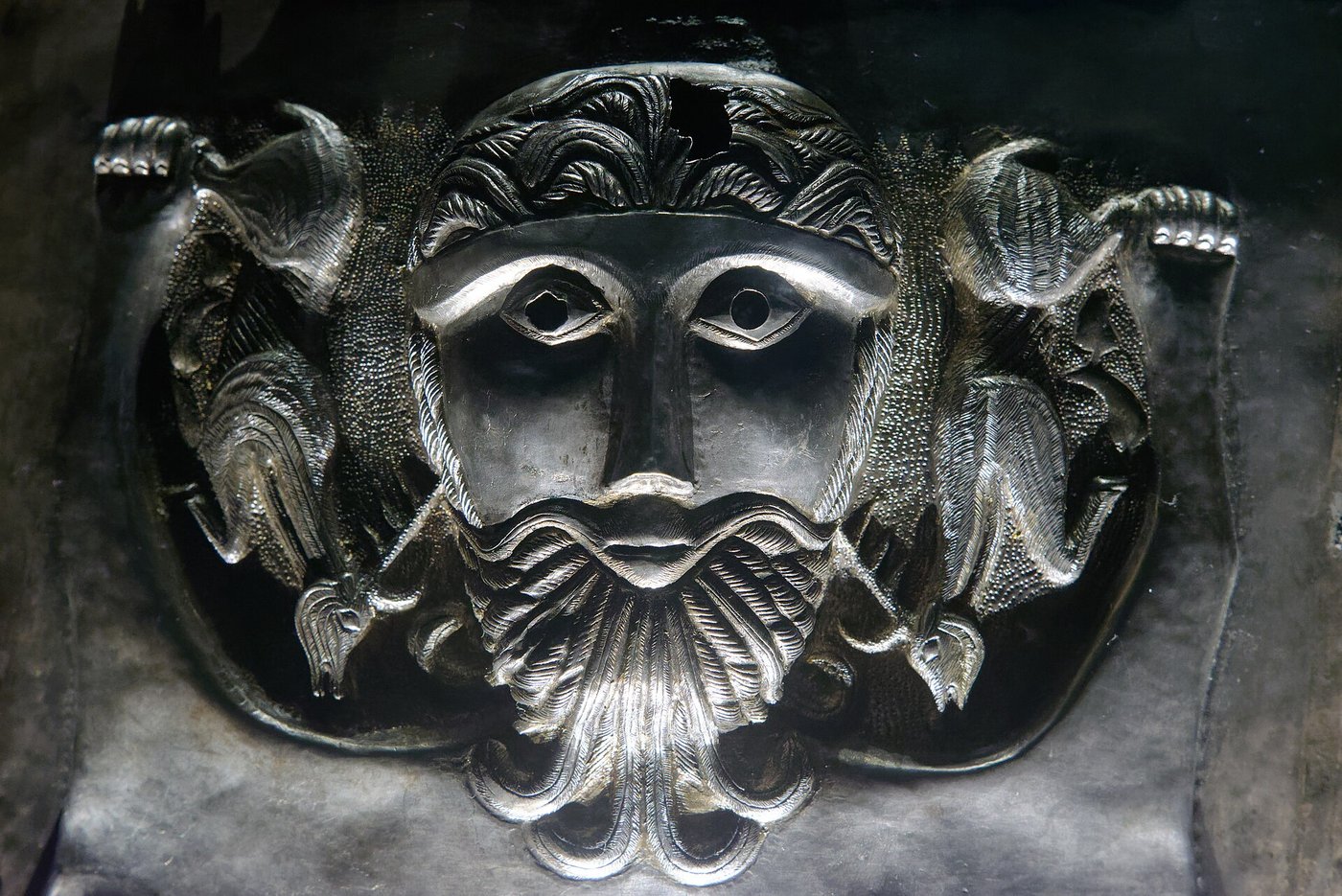 A large bearded silver face in high relief on an outer plate of the Gundestrup cauldron, with small figures worked into the hair on either side