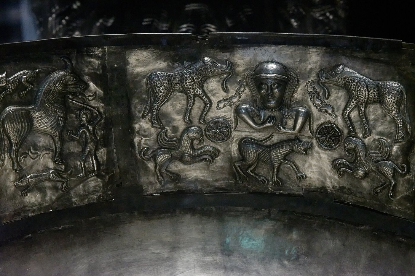 An inner plate of the Gundestrup cauldron showing a female bust flanked by spoked wheels, with two large-eared beasts resembling elephants above and rows of lions and hounds around her