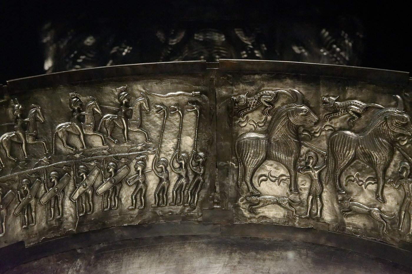 An inner plate of the Gundestrup cauldron showing marching foot-soldiers with shields below, horsemen above, three men blowing animal-headed carnyx trumpets, and a large figure lowering a smaller man head-first into a vat