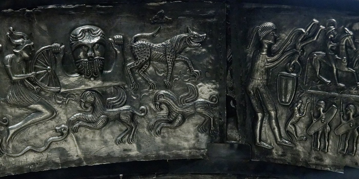 An inner plate of the Gundestrup cauldron showing a large bearded bust holding a broken wheel, with a smaller leaping figure and griffins beside a procession of horsemen
