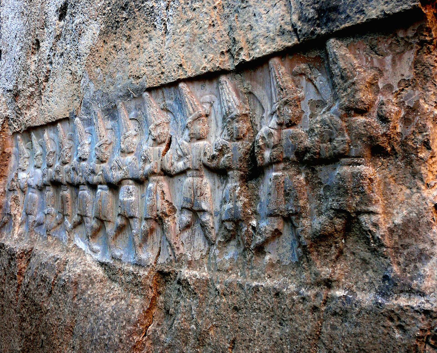 A weathered rock relief showing a row of twelve identical gods marching in step, each with a sickle-sword over one shoulder