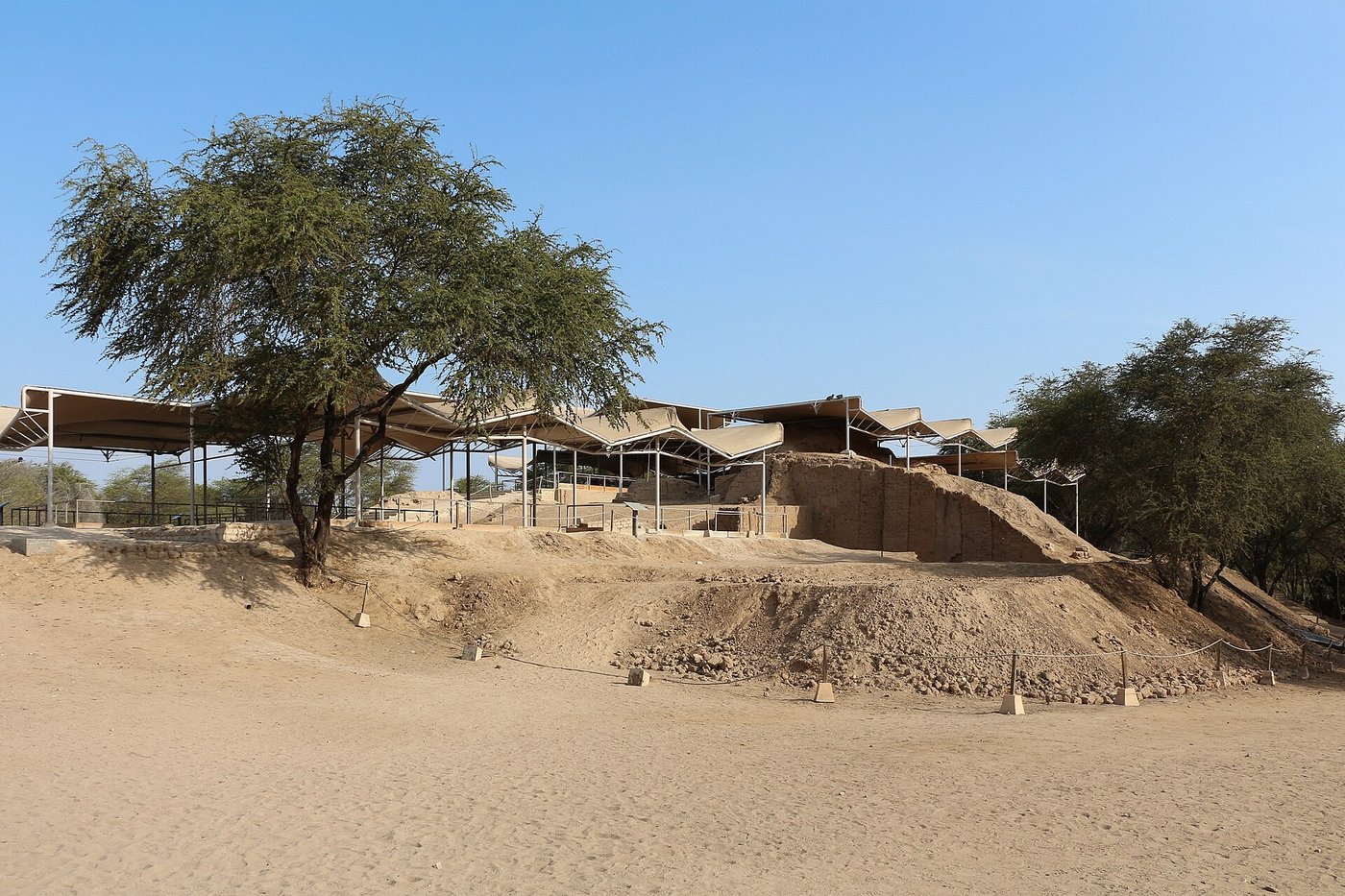 The eroded adobe pyramids of Huaca Rajada under modern shade canopies, standing in dry scrubland near Sipán, Peru