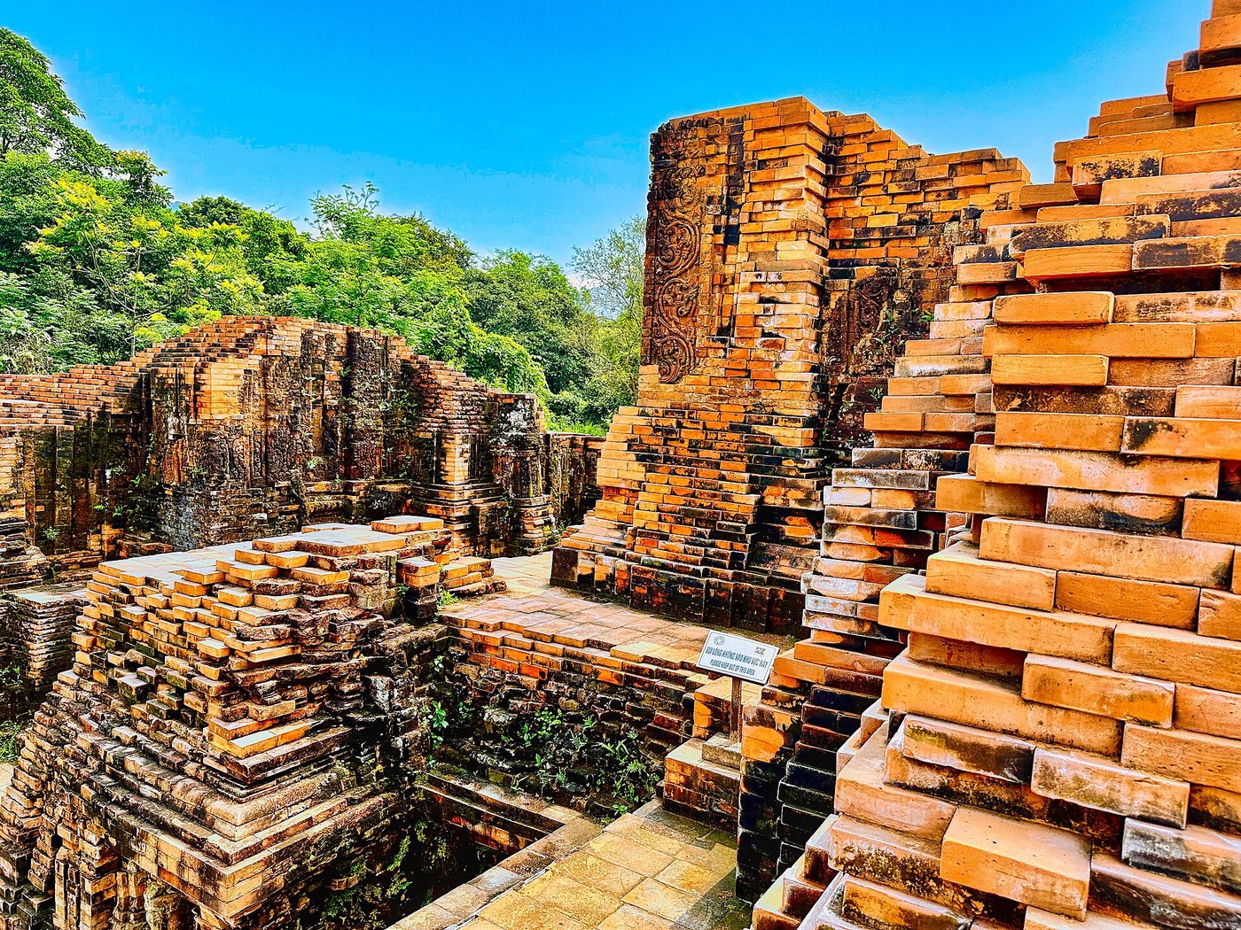 Ruined brick structures of My Son Group A among grass and trees, low walls and rubble
