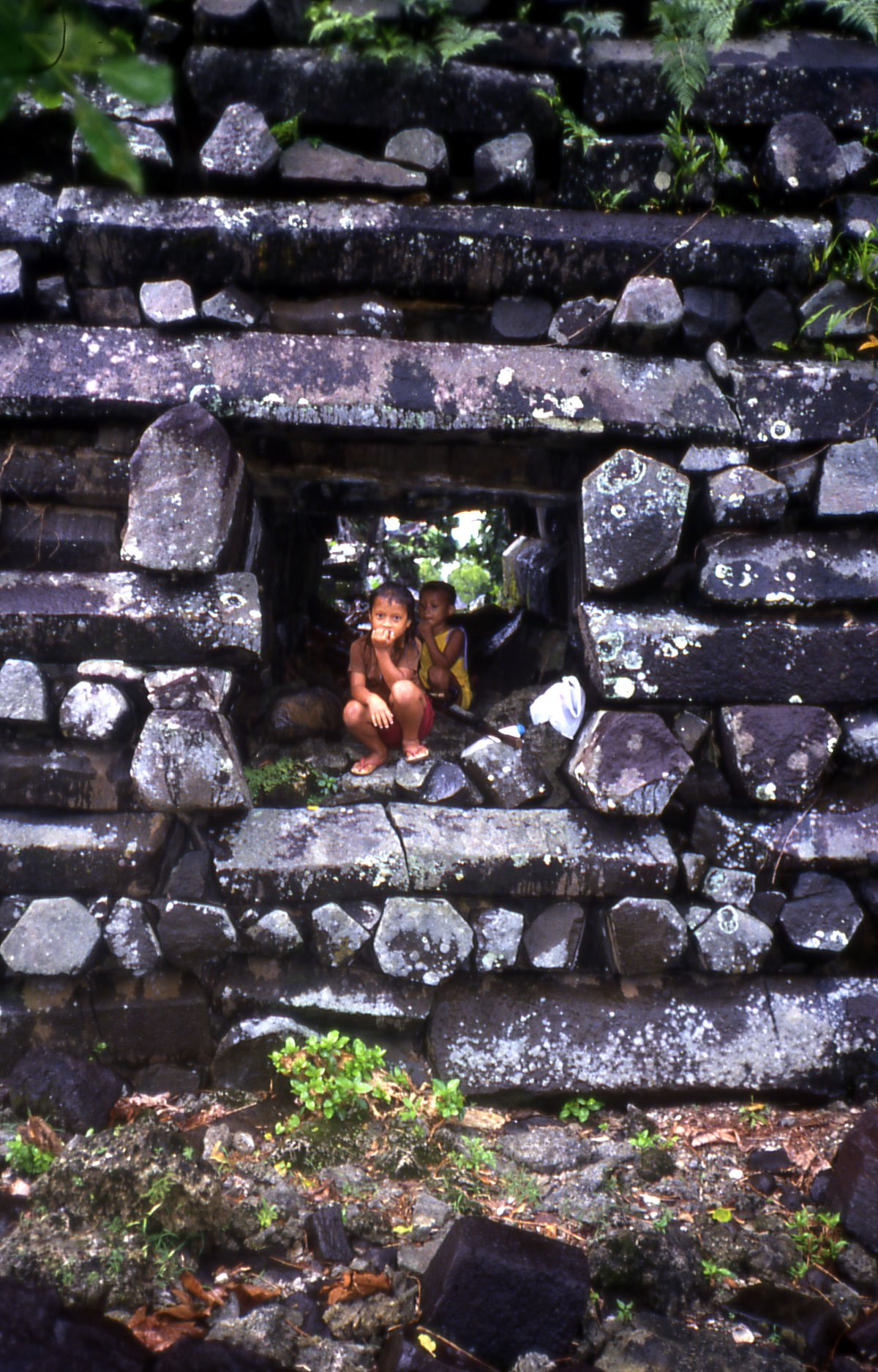 Two children standing in a narrow passage between towering basalt walls at Nan Madol, showing the scale of the stones