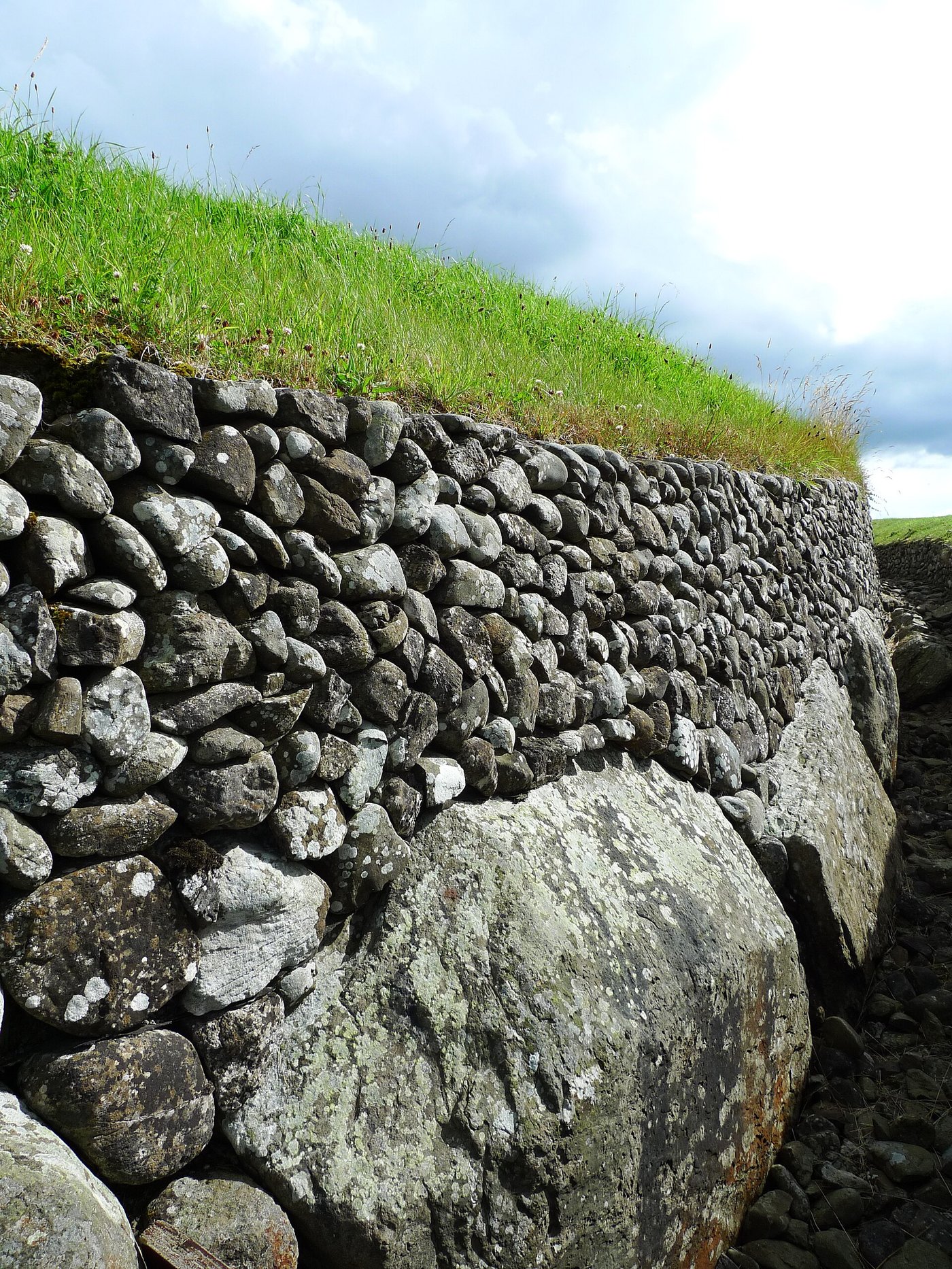 A massive carved kerbstone at the base of Newgrange with rounded cobbles and the grassed dome of the mound rising above it