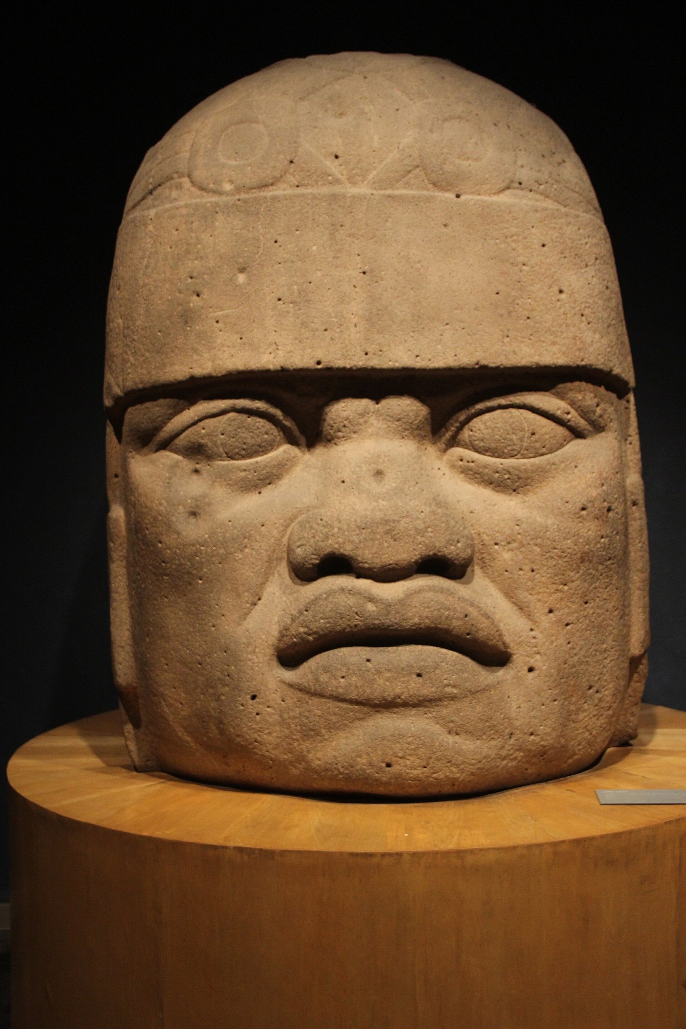 A colossal Olmec basalt head with a helmet-like headdress and heavy features, seen from the front in a museum park.