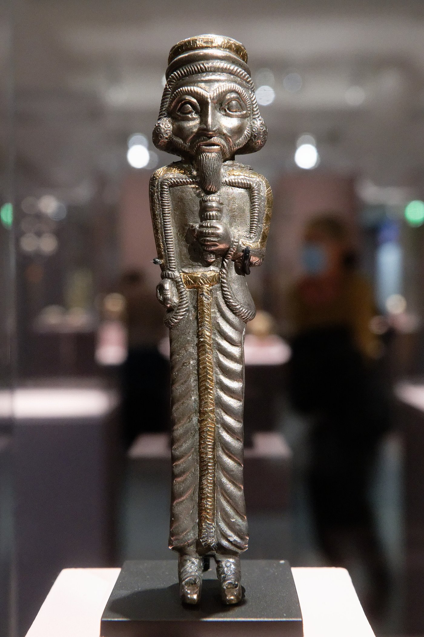 Tall thin gold figure of a standing man in Persian dress, holding a bundle of thin rods in front of him