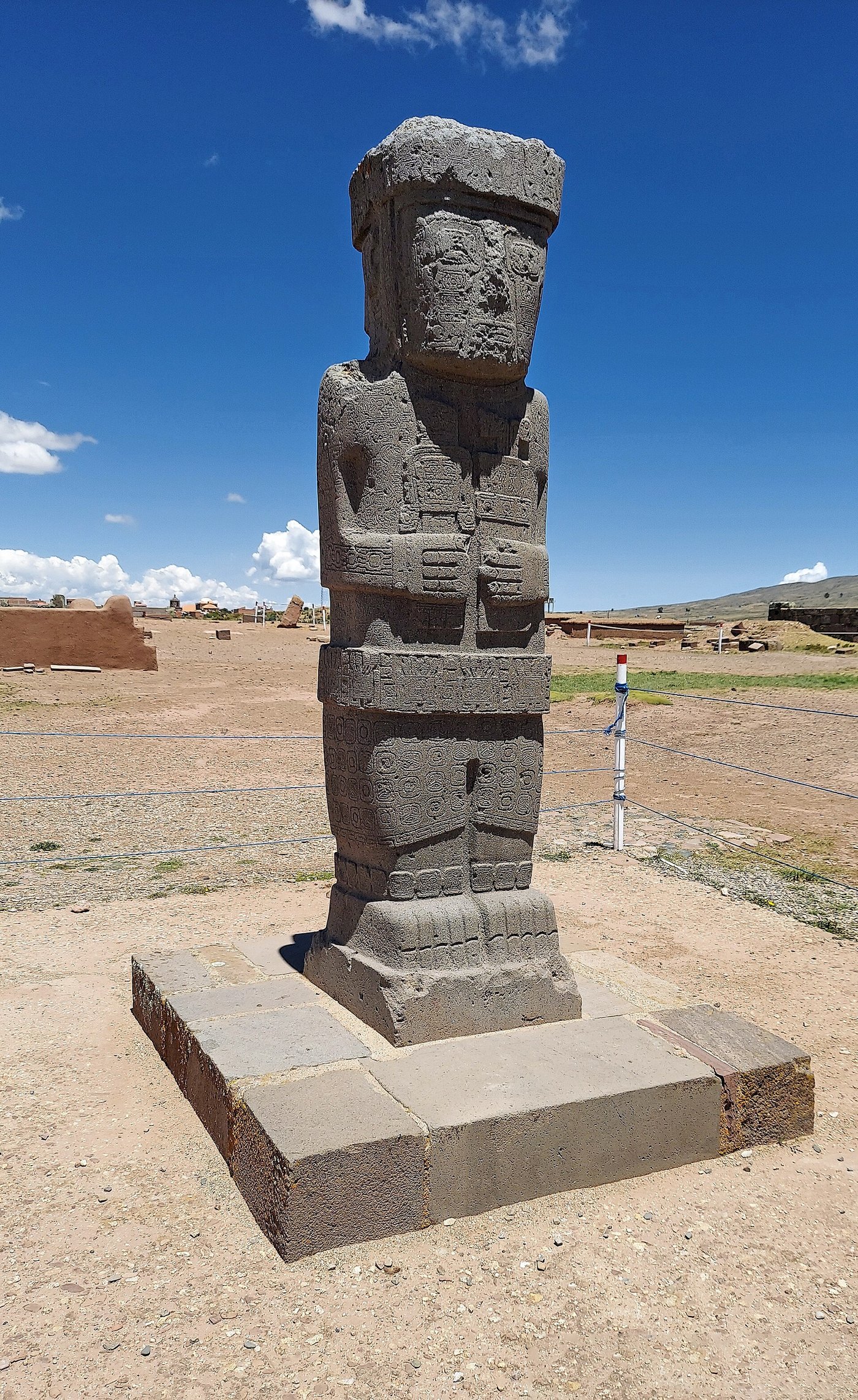 A tall squared stone statue at Tiwanaku, a stylised human figure holding a cup and a tablet, its surface covered in carving